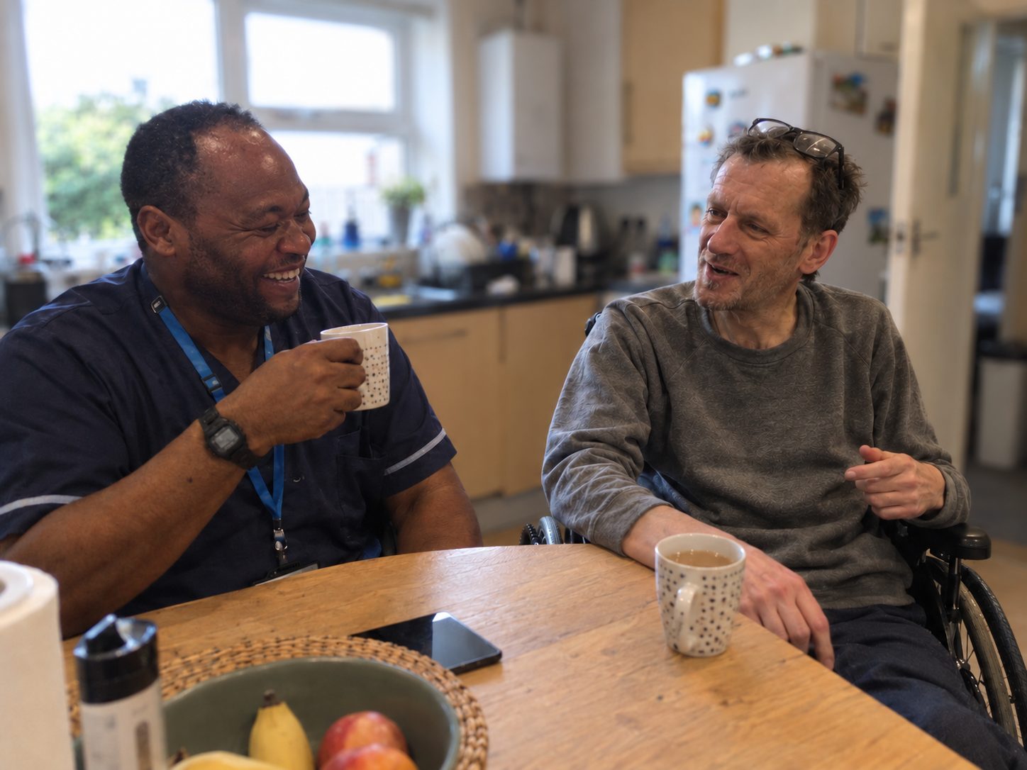 A nurse chats over tea with a man in a wheelchair at his kitchen table